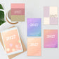 Five pastel 2027 calendar cover designs are arranged on a light gray surface with a tablet, mug, and green leaves as styled props. The covers feature soft color-blocked and gradient designs in pink, peach, lavender, purple, and beige, with the year '2027' printed prominently.