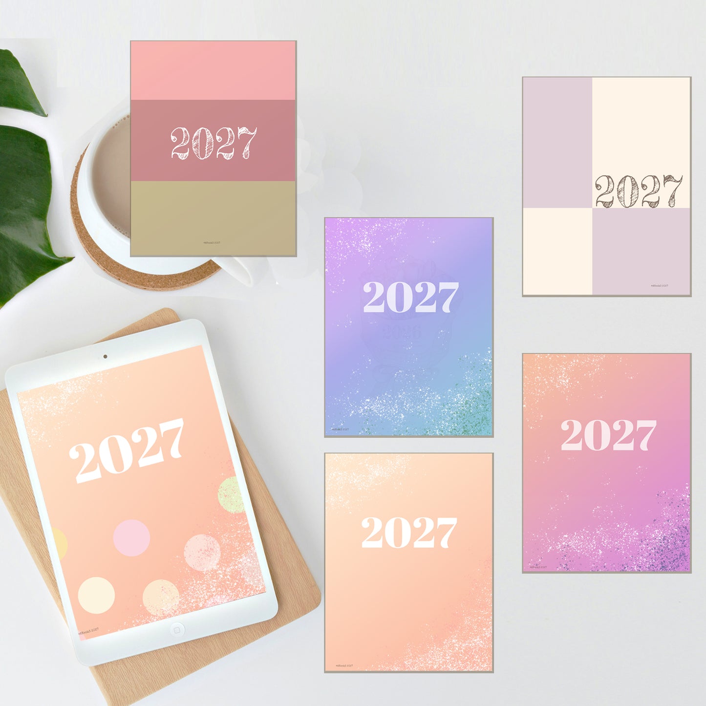 Five pastel 2027 calendar cover designs are arranged on a light gray surface with a tablet, mug, and green leaves as styled props. The covers feature soft color-blocked and gradient designs in pink, peach, lavender, purple, and beige, with the year '2027' printed prominently.